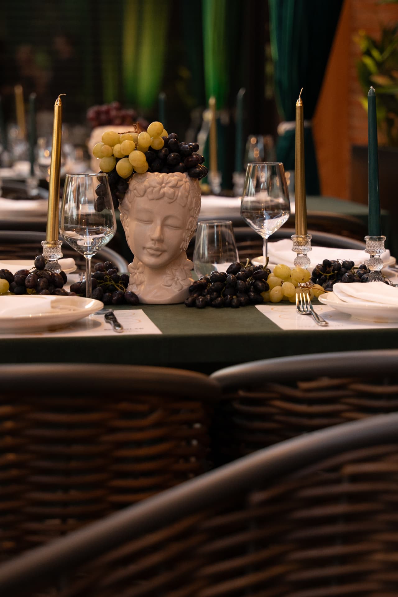 Table laid with grapes, gold and green candles and wicker chairs in front of ASADOR's green curtains