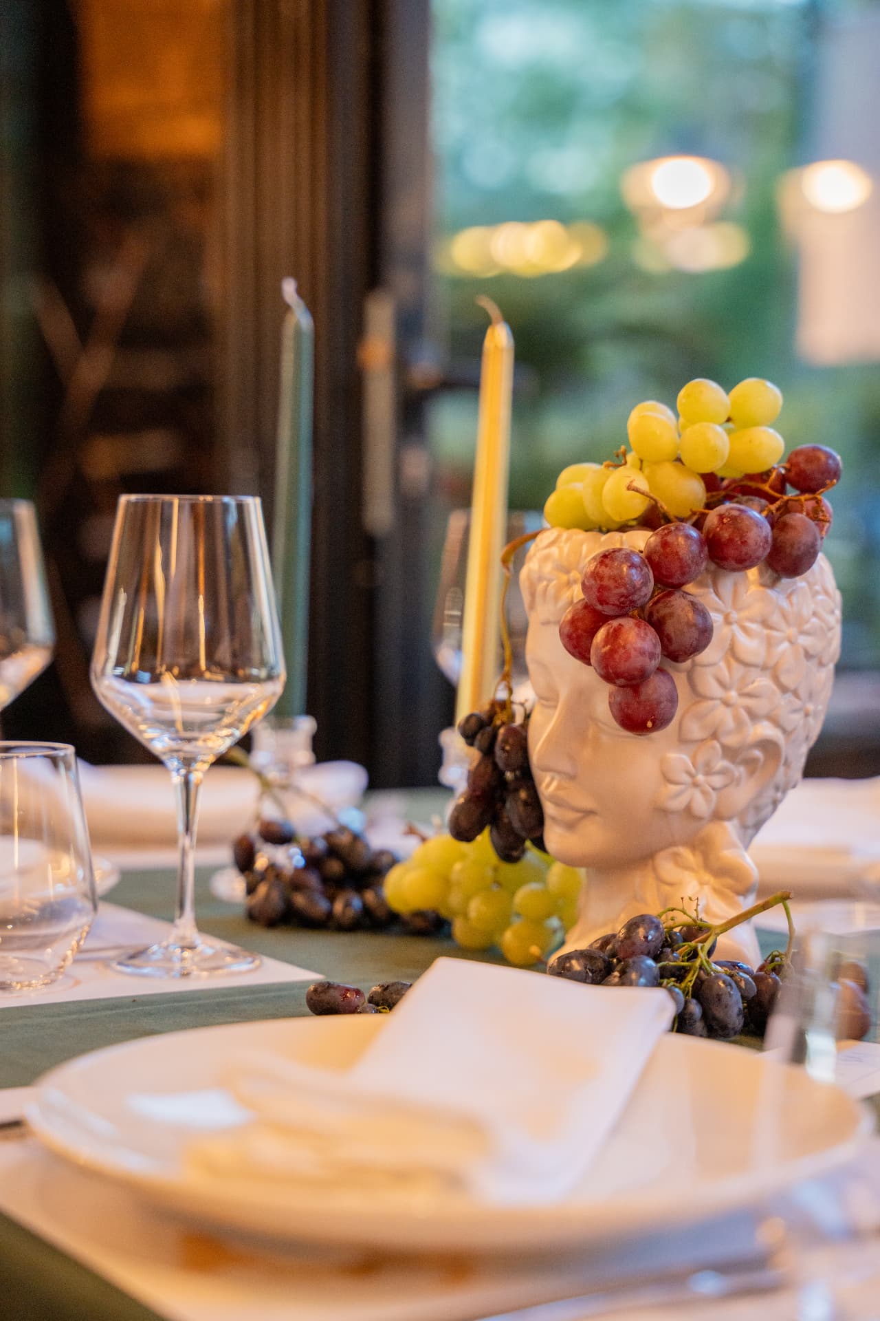 Close view of a white ceramic head vase with grapes, candles and crystal glassware on a green table runner