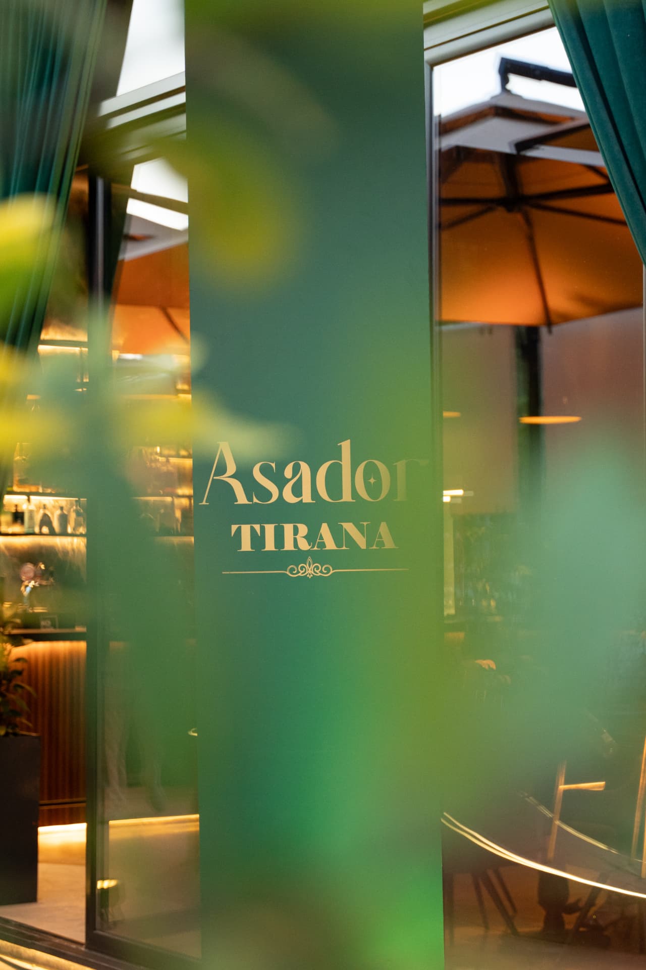 The Asador Tirana name in gold on the green glass entrance, with the softly lit interior behind
